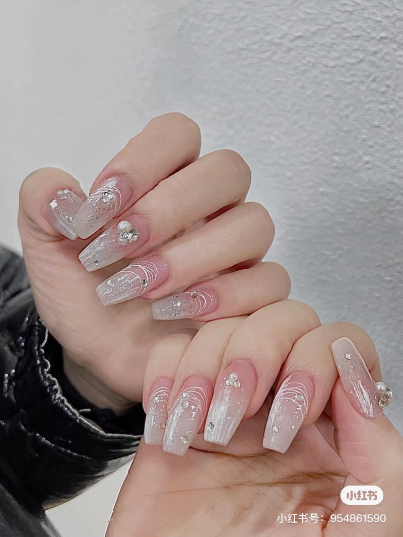 Bộ móng tay được sơn màu hồng nhạt, trang trí hoa văn trắng và đính đá lấp lánh.