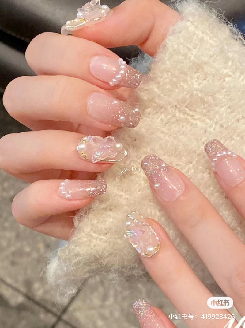 Bàn tay với móng tay đính đá và ngọc trai, thiết kế lấp lánh, nền lông màu kem.