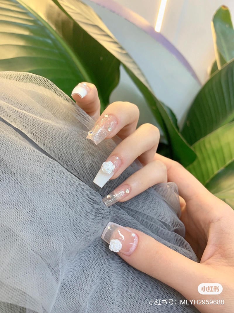 Bàn tay với móng tay được trang trí hoa và kim tuyến, đặt trên vải voan xám, nền là lá cây xanh.