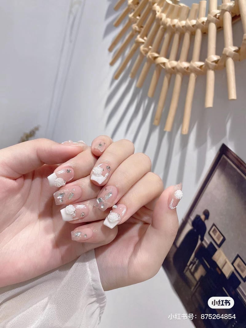 Bàn tay với móng tay được trang trí hoa trắng và kim tuyến, nền có gương và bức tranh treo tường.