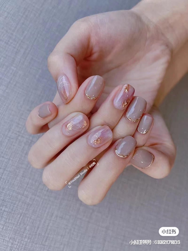 Bàn tay với móng tay được sơn màu nude, trang trí họa tiết vàng và trắng trên nền vải xám.