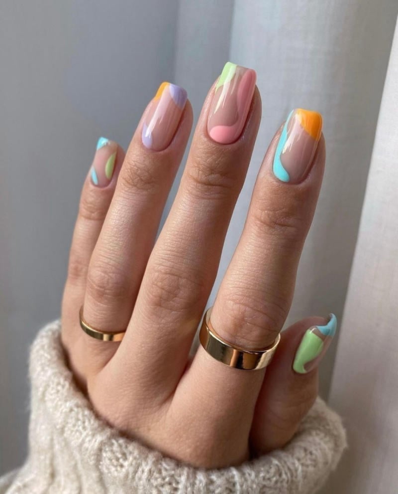 Bàn tay với móng tay sơn nhiều màu sắc pastel, đeo nhẫn vàng trên hai ngón, nền trắng mờ.