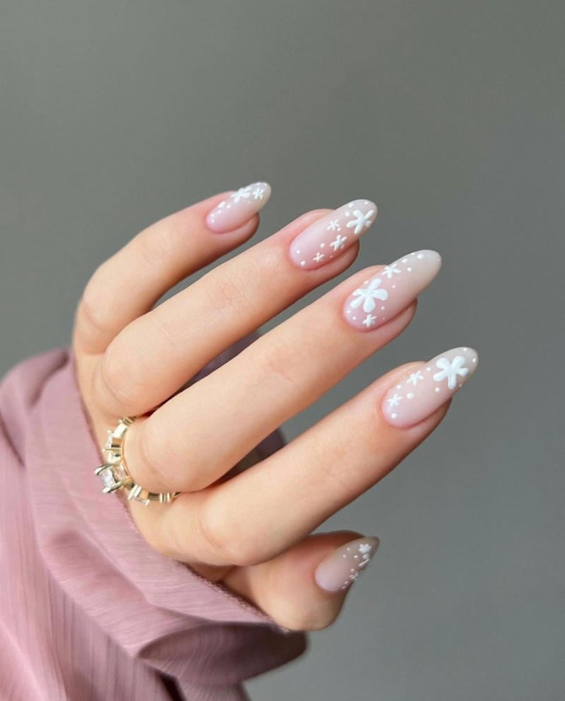 Bàn tay đeo nhẫn, móng tay sơn màu hồng nhạt với họa tiết hoa trắng và áo tay dài màu hồng nhạt.
