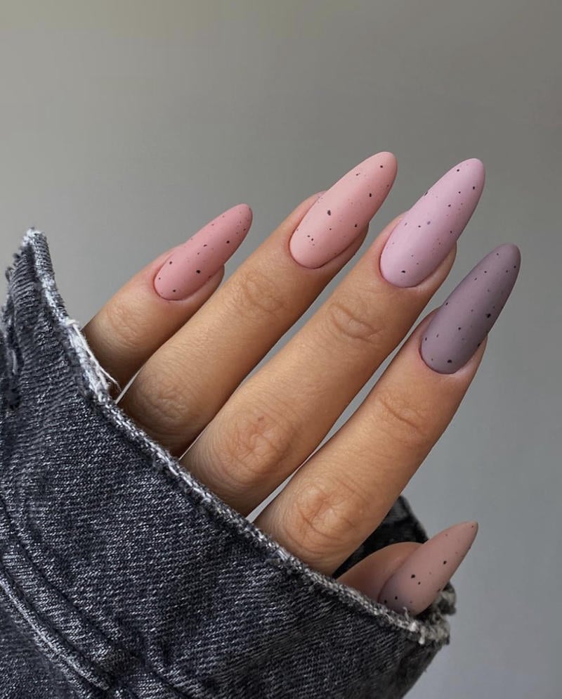 Bàn tay với móng tay dài, sơn màu hồng và tím nhạt, có chấm đen, áo khoác denim xám.