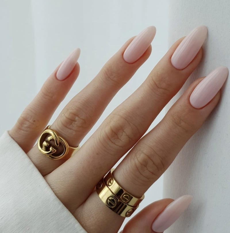 Bàn tay với móng tay dài màu hồng nhạt, đeo ba chiếc nhẫn vàng trên ngón tay giữa và áp út.