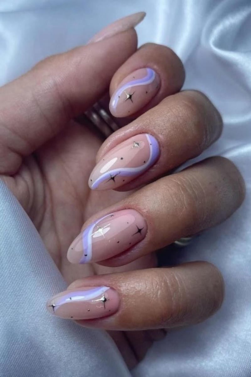 Bàn tay với móng tay được sơn màu nude, trang trí họa tiết lượn sóng tím và điểm nhấn ánh sao trên nền vải trắng.