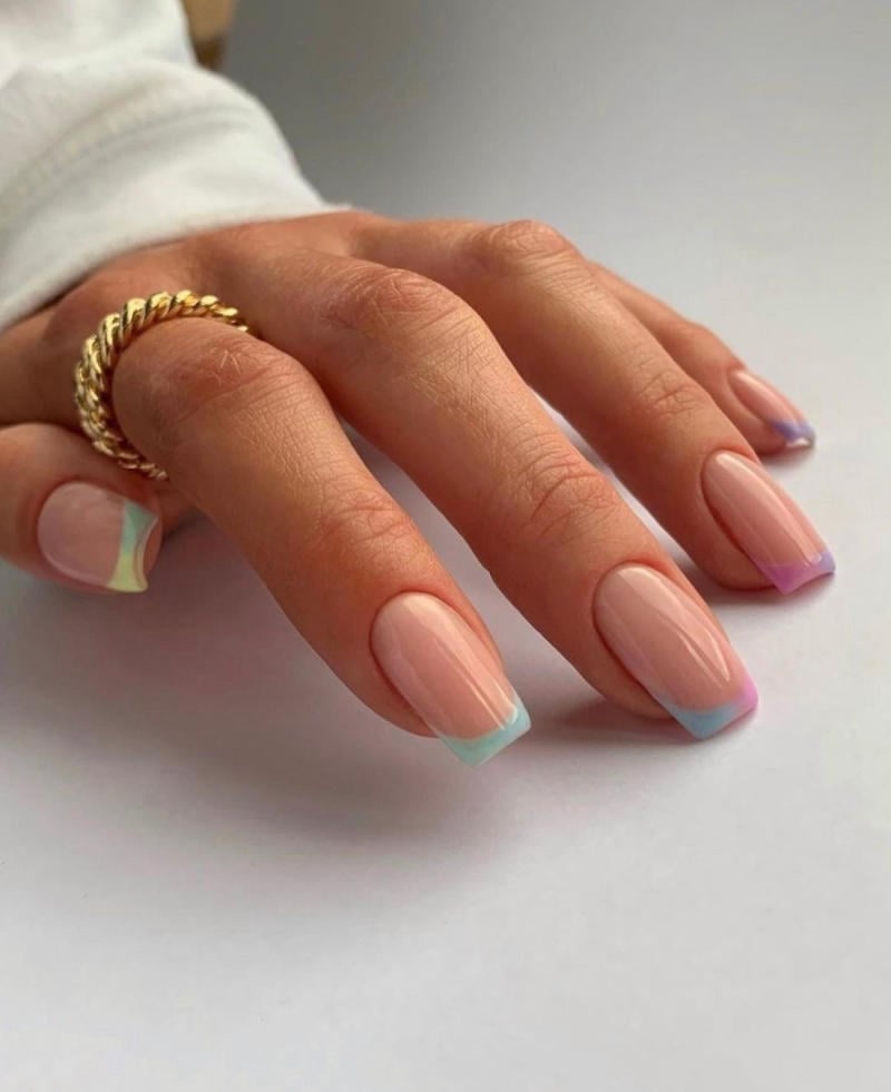Bàn tay đeo nhẫn vàng, móng tay sơn màu pastel với viền đầu móng nhiều màu sắc trên nền trắng.