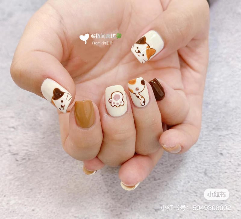 Bàn tay với móng tay trang trí hình mèo và các họa tiết màu nâu, trắng, đen trên nền màu be.