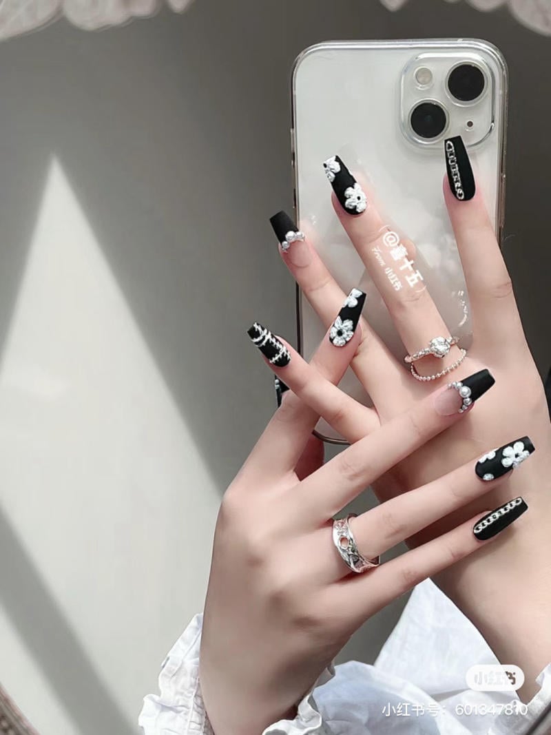 Bàn tay với móng tay đen trang trí hoa trắng, cầm điện thoại, đeo nhẫn bạc lấp lánh, ánh sáng chiếu nhẹ.