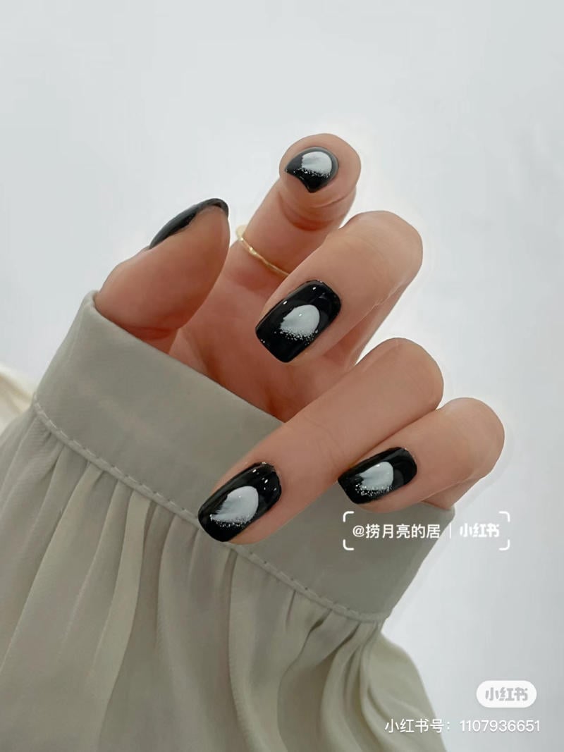 Bàn tay với móng tay sơn màu đen, họa tiết trăng trắng, đeo nhẫn vàng, trên nền áo màu xám nhạt.