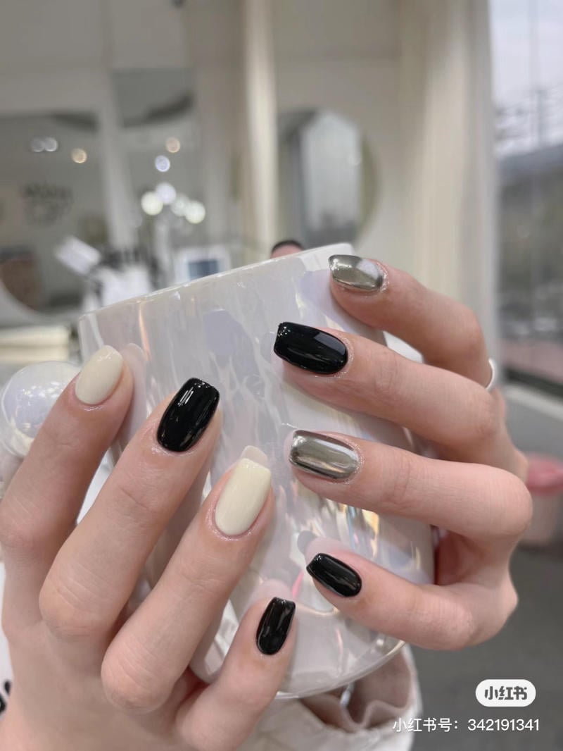 Bàn tay với móng tay được sơn màu đen, trắng và bạc, đang cầm một chiếc cốc trắng có hoa văn.