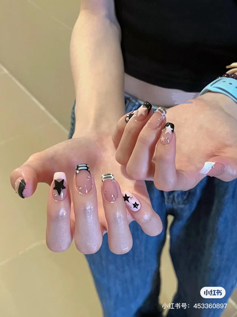 Bàn tay với móng tay được trang trí họa tiết ngôi sao màu đen và sọc trắng trên nền màu hồng nhạt.