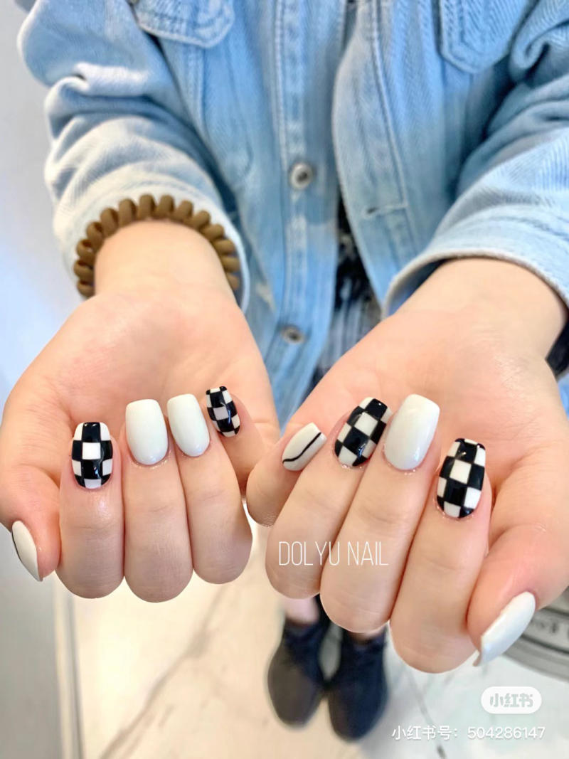 Hai bàn tay với móng tay được trang trí họa tiết caro đen trắng và sơn màu trắng, người mặc áo khoác xanh.