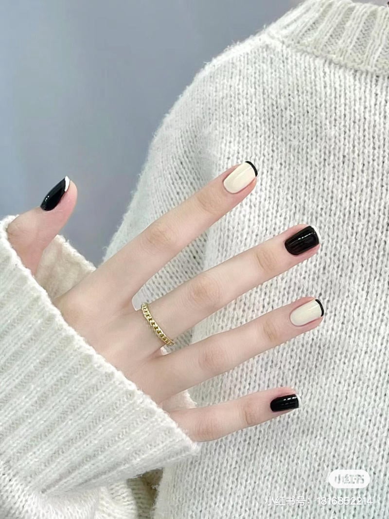 Một bàn tay đeo nhẫn vàng với móng tay sơn đen và trắng, mặc áo len màu kem mềm mại.