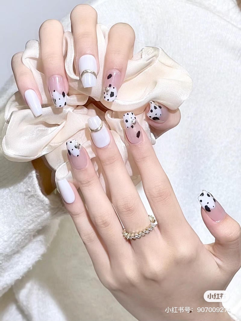 Bàn tay với móng tay sơn trắng đốm đen, đeo nhẫn vàng, đặt trên nền vải lụa trắng.