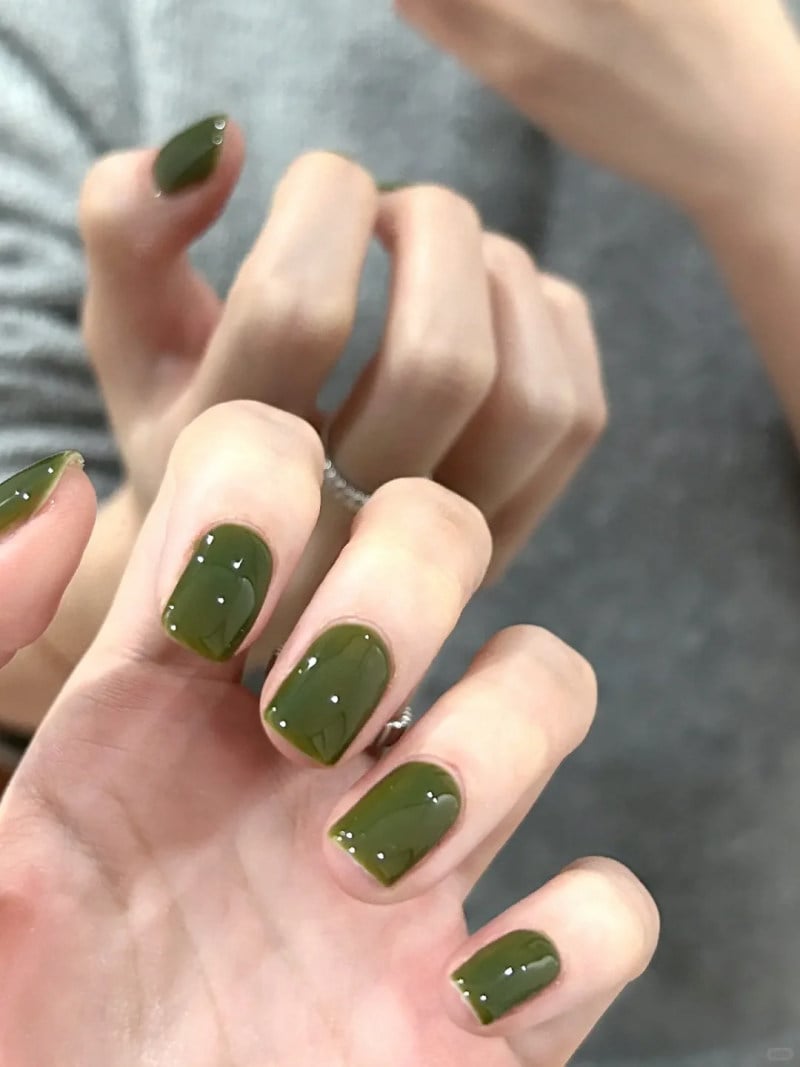 B&agrave;n tay với m&oacute;ng tay sơn m&agrave;u xanh &ocirc; liu, đang tạo d&aacute;ng trước gương, nền &aacute;o x&aacute;m.