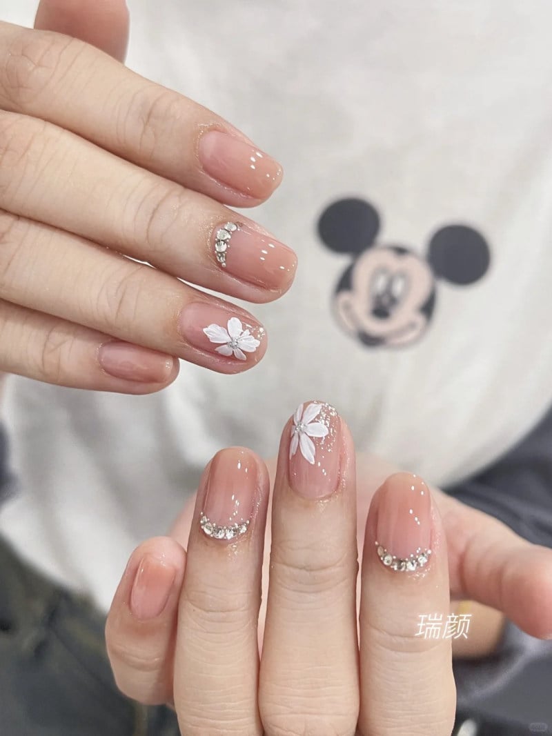 B&agrave;n tay với m&oacute;ng tay được sơn m&agrave;u nude, trang tr&iacute; hoa trắng v&agrave; đ&iacute;nh đ&aacute; nhỏ, nền &aacute;o in h&igrave;nh nh&acirc;n vật hoạt h&igrave;nh.