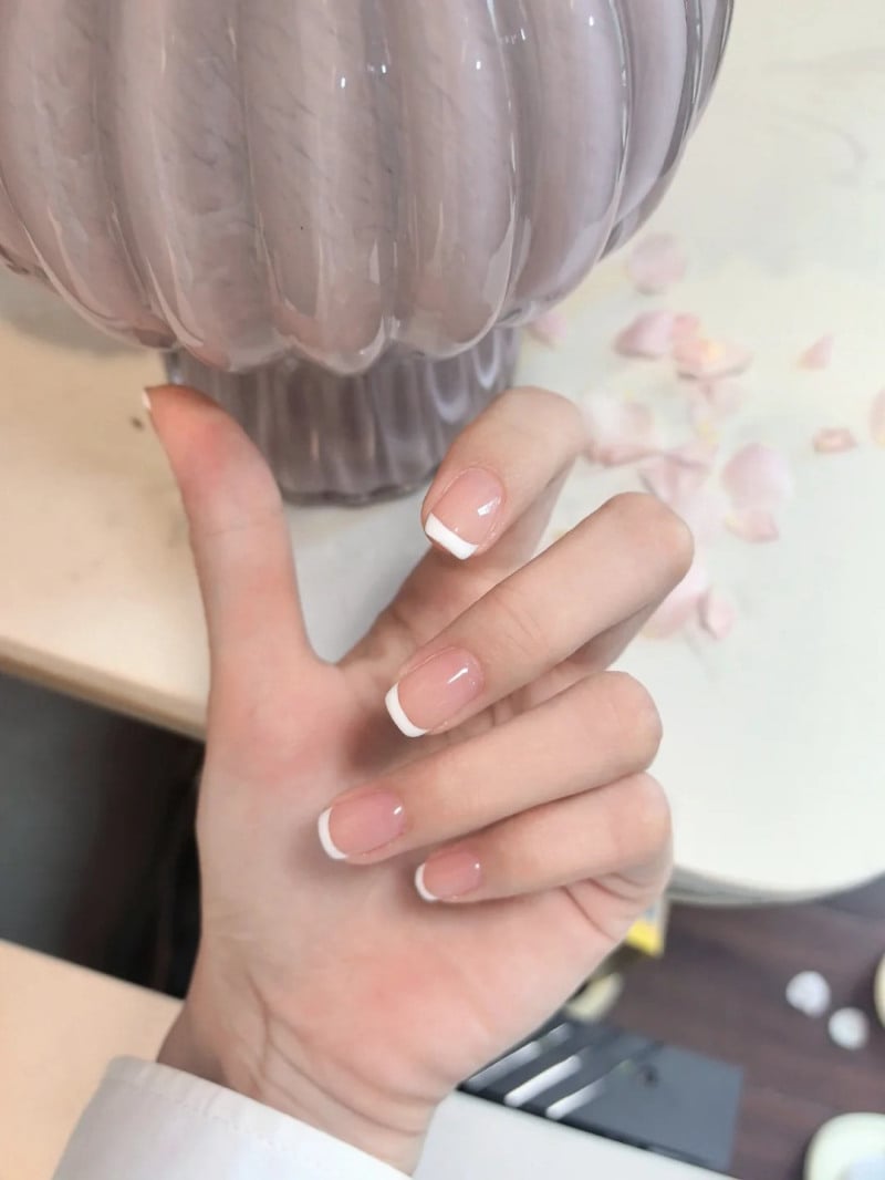 Bàn tay với móng tay kiểu Pháp cầm chậu cây màu tím nhạt, trên bàn có vài cánh hoa rơi.