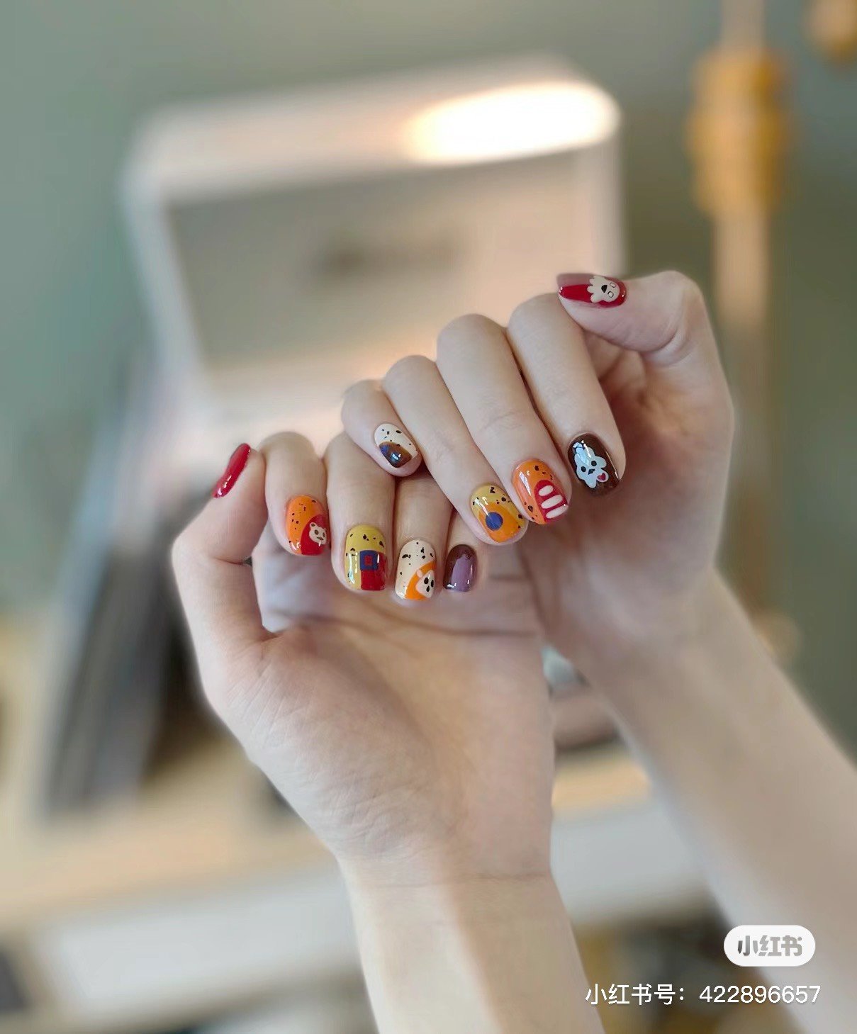 Bàn tay với móng được trang trí nghệ thuật, nhiều màu sắc và họa tiết đa dạng, chụp trong không gian nội thất.