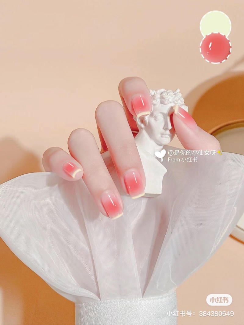 Bàn tay với móng tay ombre đỏ hồng cầm tượng nhỏ màu trắng, đặt trên nền vải trắng.
