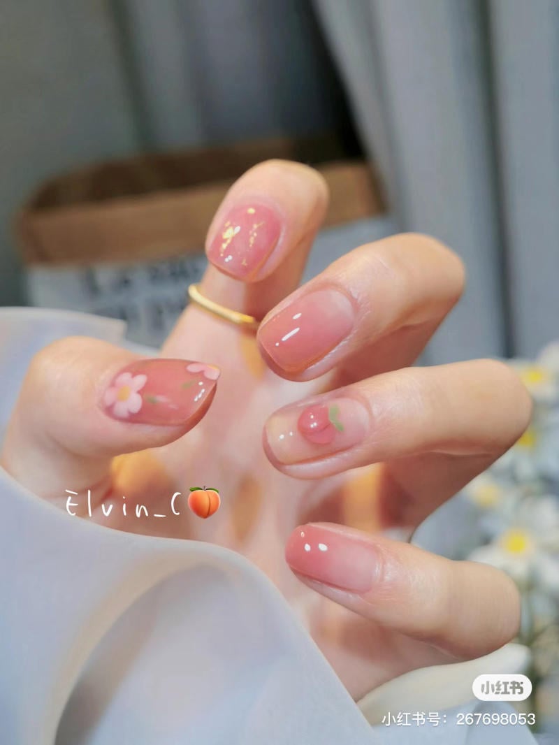 Bàn tay với móng tay trang trí hoa và hoạ tiết trái cây màu hồng nhạt, đeo nhẫn vàng.