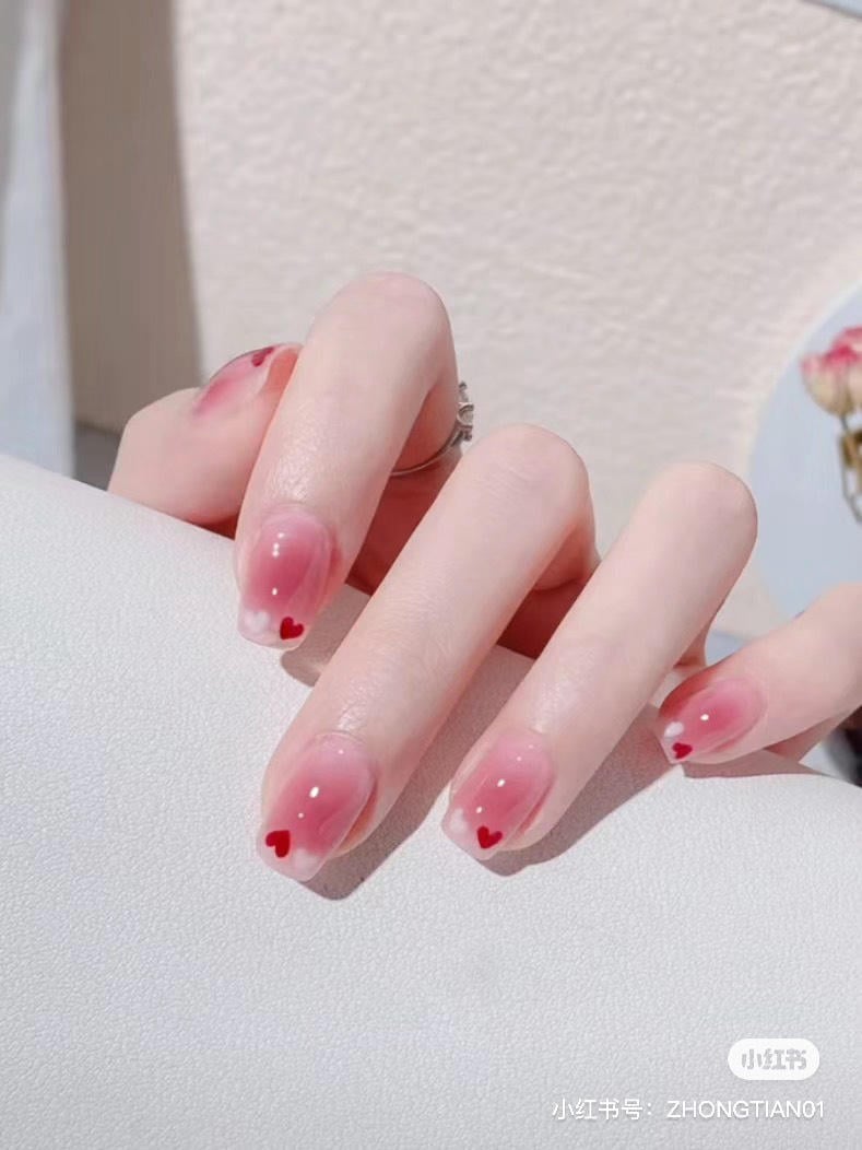 Bàn tay với móng tay sơn màu hồng nhạt, có họa tiết trái tim đỏ, đặt trên bề mặt sáng màu.