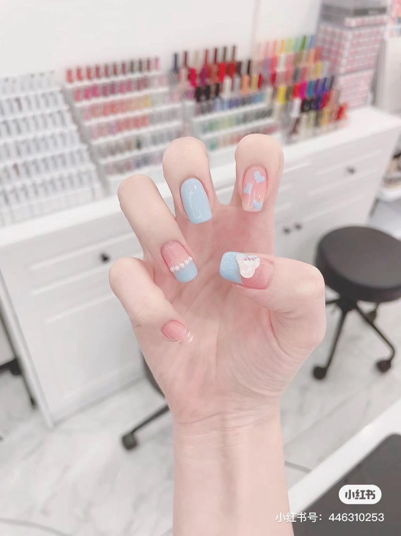 Bàn tay với móng tay được sơn màu xanh nhạt và hồng, có họa tiết, trong tiệm làm móng với kệ sơn nhiều màu.