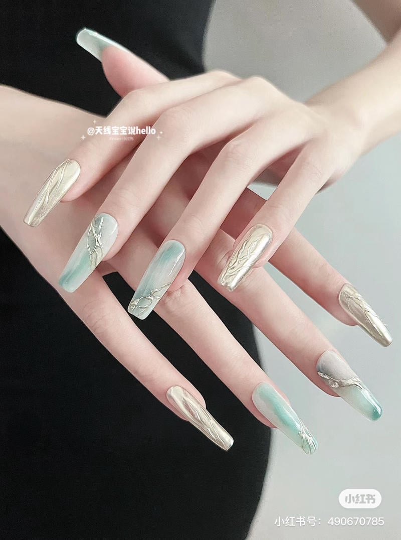 Bàn tay với móng tay dài được sơn màu xanh và vàng ánh kim, thiết kế họa tiết trang trí tinh tế.