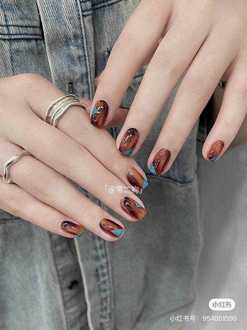 Bàn tay với móng tay sơn màu nâu và xanh, đeo nhẫn bạc, áo khoác denim nền.