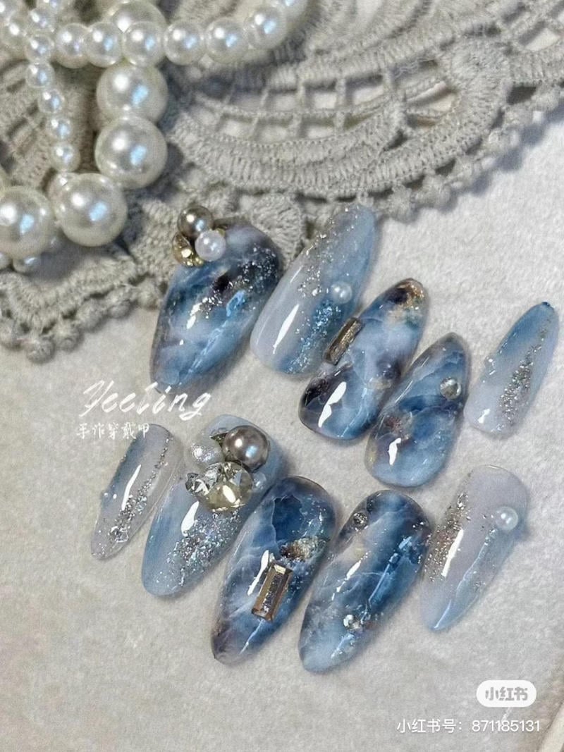 Bộ móng tay giả màu xanh với trang trí ngọc trai và kim cương giả, đặt trên nền vải ren.