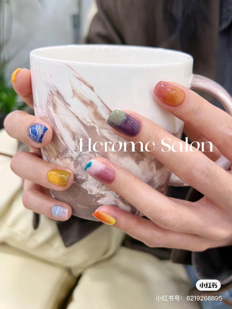 Tay cầm ly, móng tay được sơn nhiều màu sắc khác nhau, nền mờ có chữ "Herome Salon".