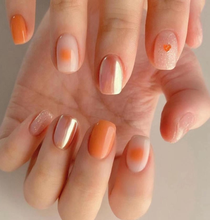 B&agrave;n tay với m&oacute;ng tay trang tr&iacute; m&agrave;u cam v&agrave; kim tuyến, c&oacute; họa tiết tr&aacute;i tim nhỏ tr&ecirc;n một m&oacute;ng.