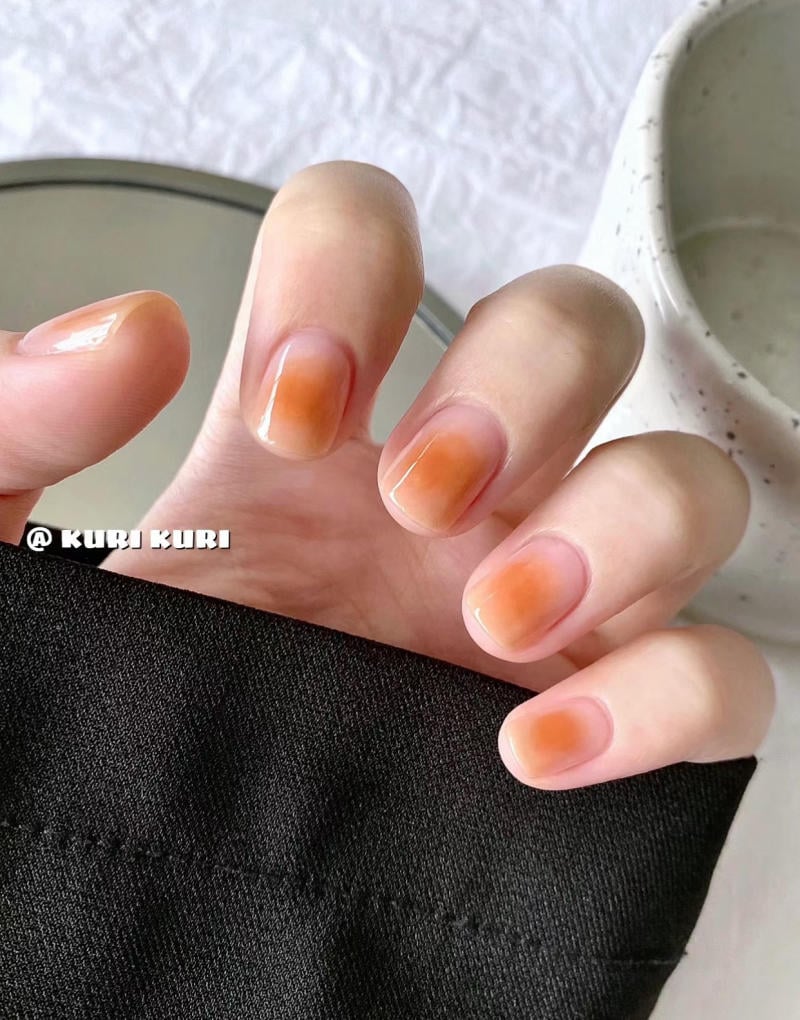 B&agrave;n tay c&oacute; m&oacute;ng tay sơn m&agrave;u cam nhạt, cầm một miếng vải m&agrave;u đen, nền c&oacute; một chiếc cốc m&agrave;u trắng.