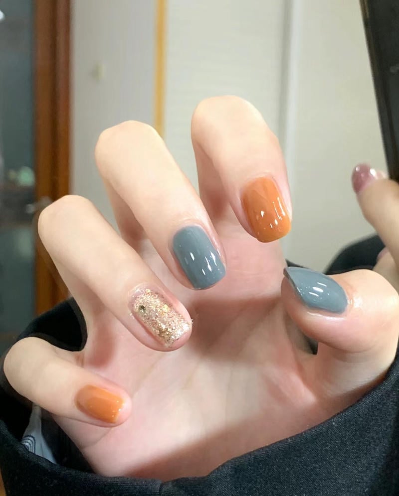 B&agrave;n tay với m&oacute;ng tay được sơn m&agrave;u cam, x&aacute;m v&agrave; nhũ v&agrave;ng, cầm một vật m&agrave;u đen.