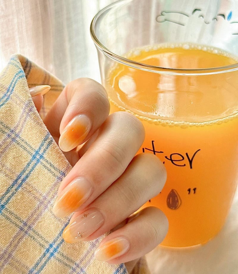 Tay cầm ly nước cam, m&oacute;ng tay sơn m&agrave;u cam nhạt, nền l&agrave; vải kẻ &ocirc; vu&ocirc;ng m&agrave;u xanh v&agrave; v&agrave;ng nhạt.