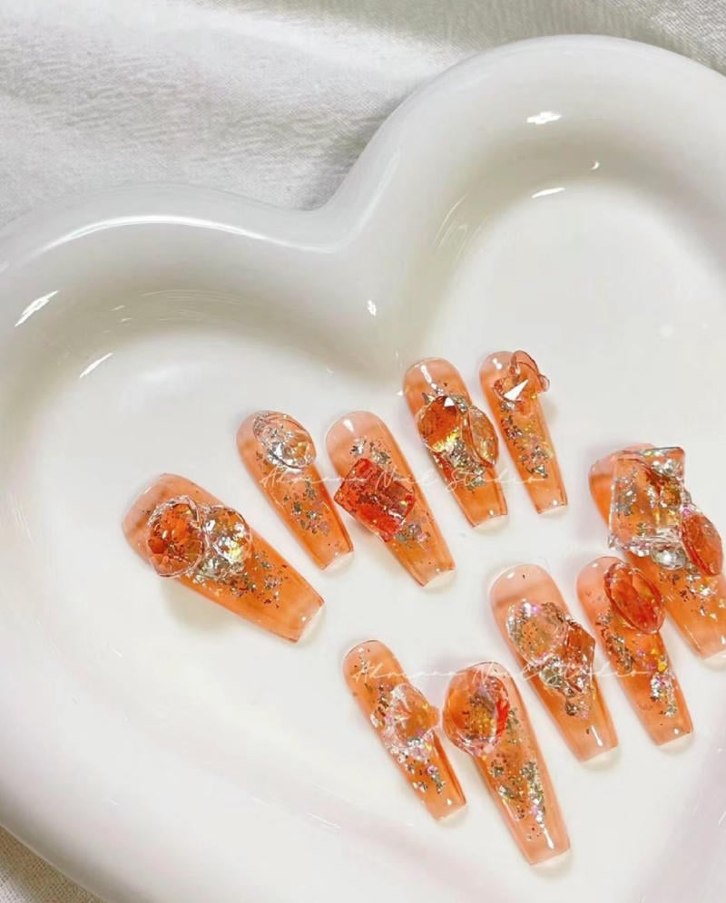 M&oacute;ng tay giả m&agrave;u cam, &aacute;nh nhũ, đặt trong đĩa h&igrave;nh tr&aacute;i tim trắng.