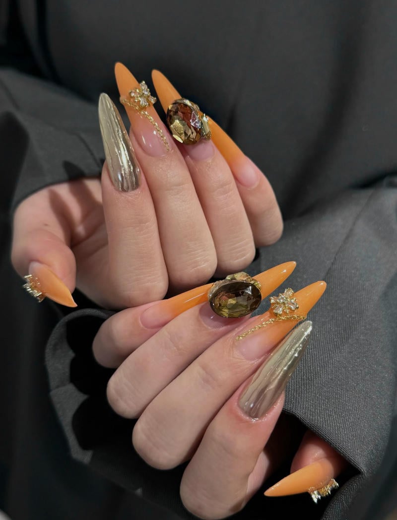 B&agrave;n tay với m&oacute;ng tay d&agrave;i, sơn m&agrave;u cam v&agrave; &aacute;nh kim loại, trang tr&iacute; đ&aacute; v&agrave; hoa nổi bật.