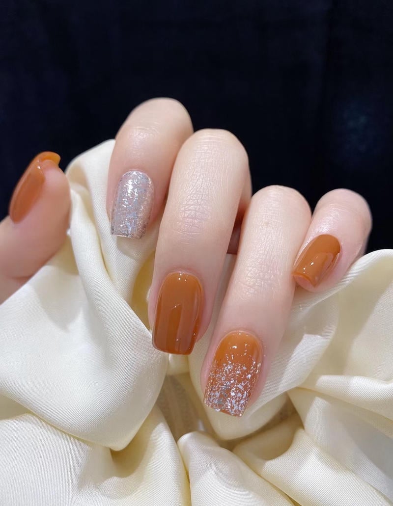 B&agrave;n tay với m&oacute;ng tay sơn m&agrave;u n&acirc;u v&agrave; bạc lấp l&aacute;nh, cầm một chiếc khăn lụa m&agrave;u kem.