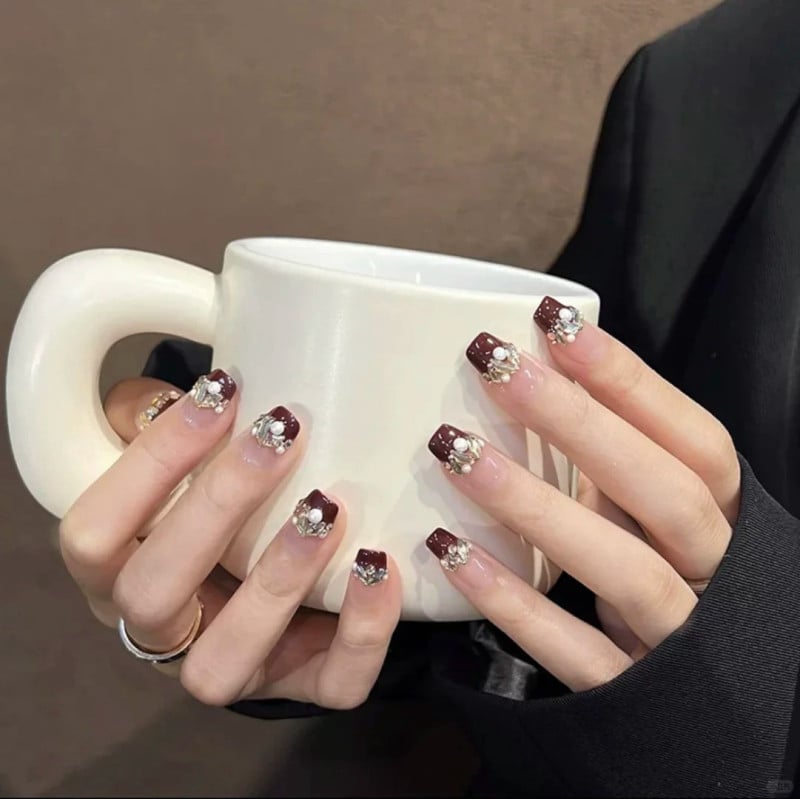 Bàn tay cầm cốc trắng, móng tay sơn màu nâu đỏ trang trí hoa và đá lấp lánh.