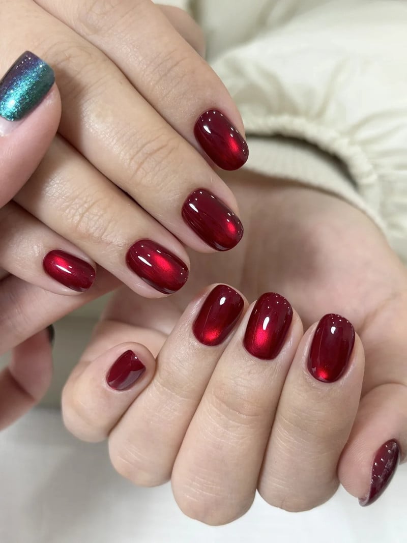 Bàn tay với móng tay sơn màu đỏ bóng và một móng sơn màu xanh lấp lánh, trên nền áo màu sáng.
