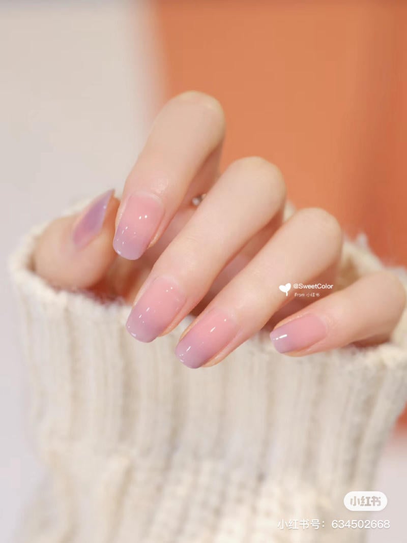 Bàn tay với móng tay được sơn màu ombre hồng tím, mặc áo len sáng màu, nền cam nhạt.