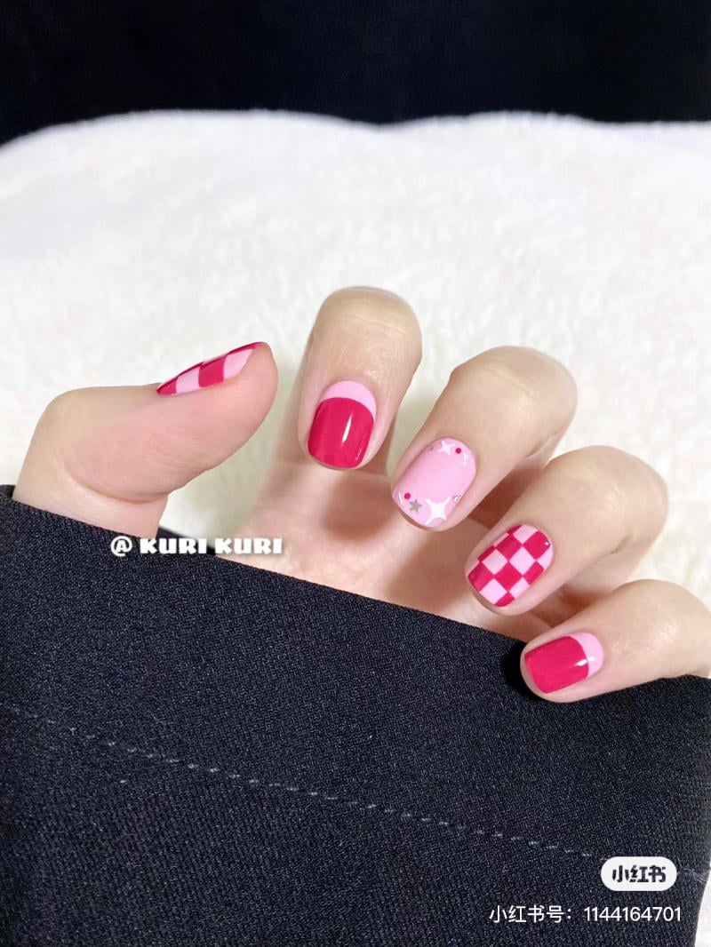 Bàn tay với móng tay được sơn màu đỏ và hồng, có họa tiết caro và ngôi sao, nền vải màu đen.