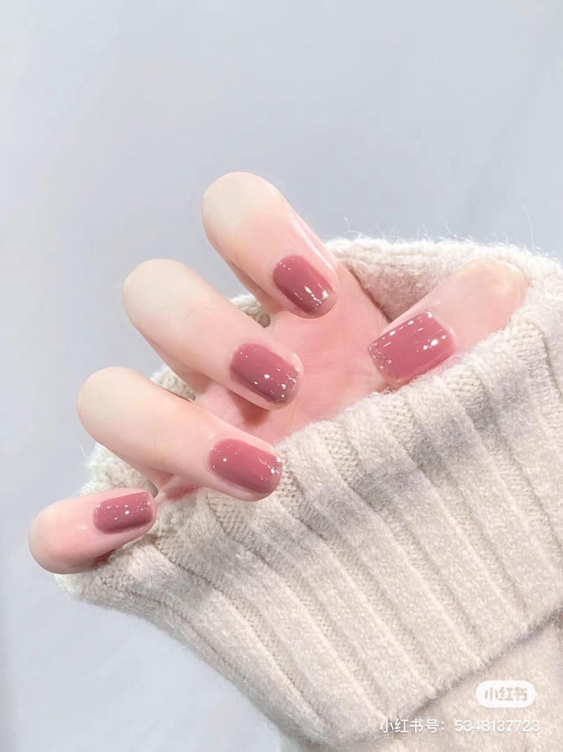 Bàn tay với móng tay sơn màu hồng, mặc áo len màu kem, nền màu xám nhạt.