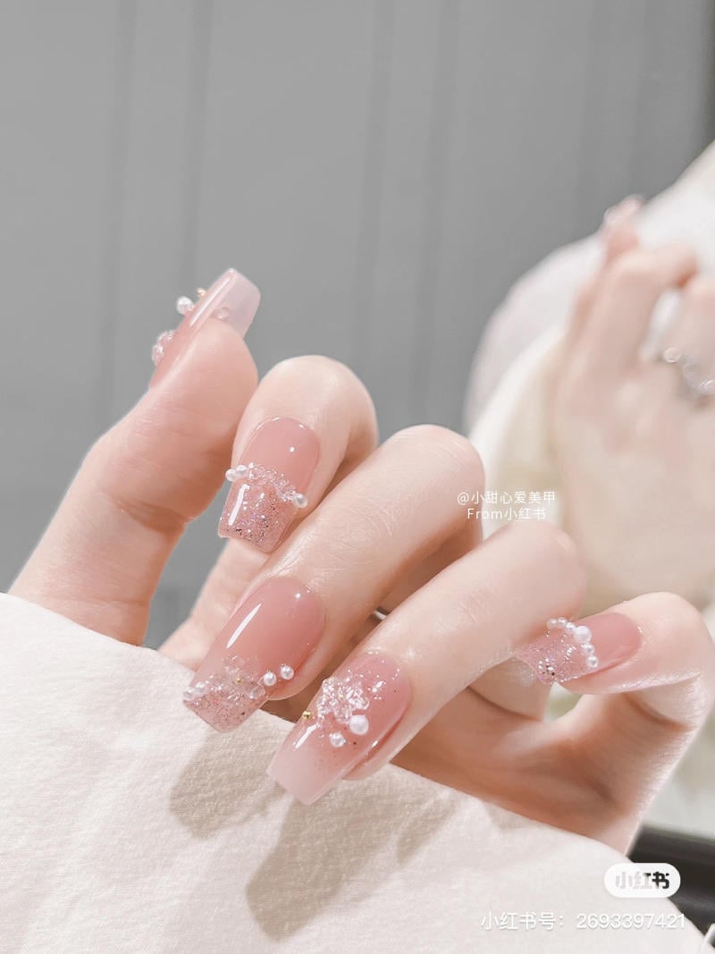 Bàn tay với móng tay dài được trang trí bằng hạt và hoa văn tinh tế trên nền màu hồng nhạt.