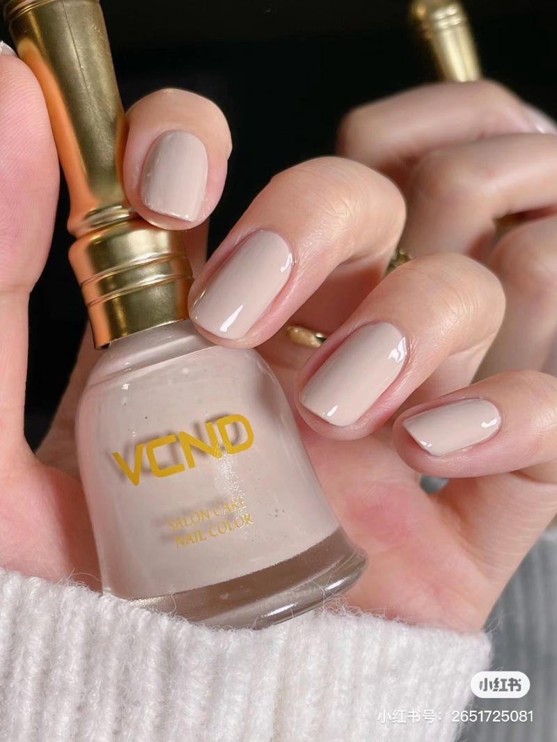 Tay cầm lọ sơn móng tay màu nude, móng tay được sơn cùng màu, nền đen, tay áo len trắng.