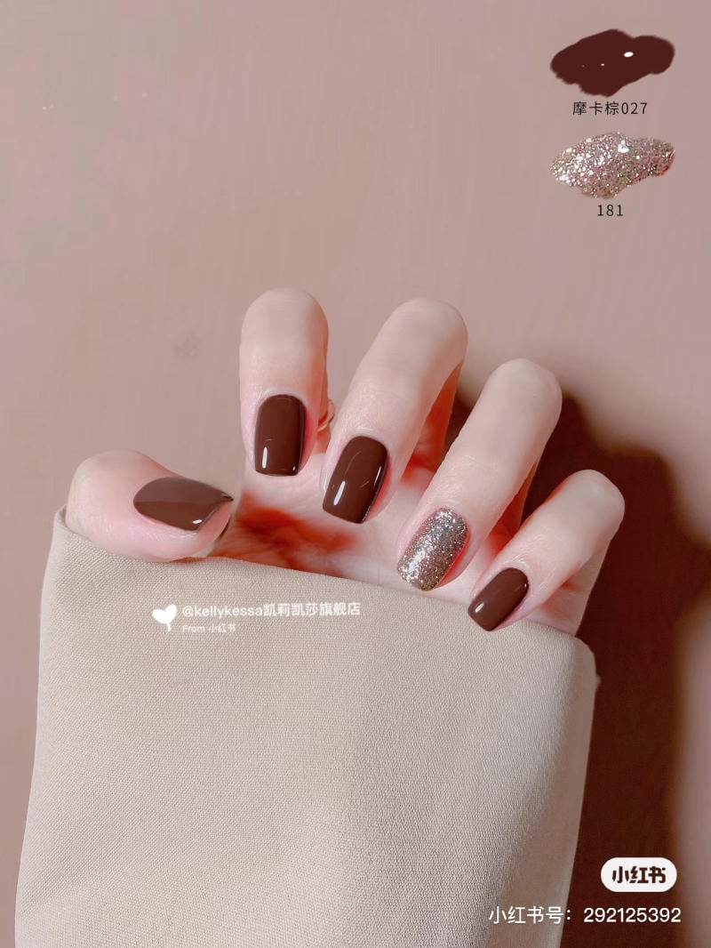 Bàn tay với móng tay sơn màu nâu và một móng nhũ bạc, nền vải màu be.
