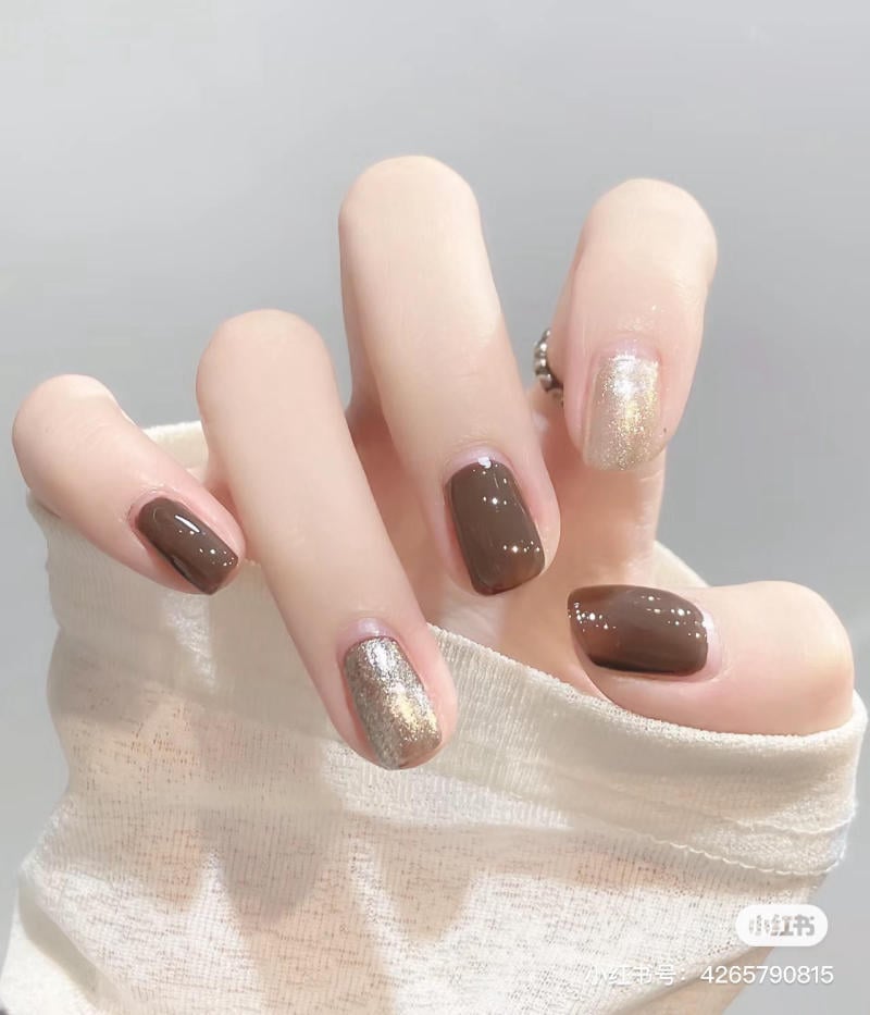 Bàn tay với móng tay sơn màu nâu và nhũ vàng lấp lánh, ngón tay đeo nhẫn, áo tay dài màu trắng.
