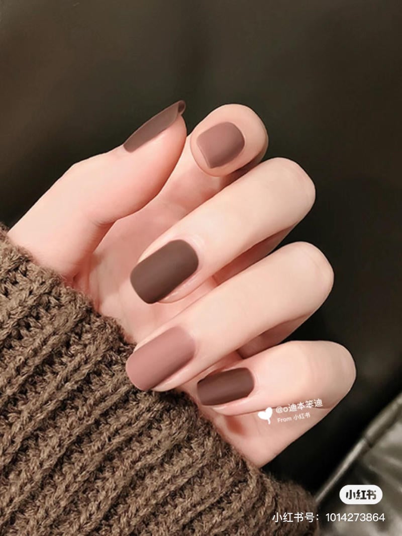 Bàn tay với móng tay sơn màu nâu mờ, mặc áo len màu nâu, nền tối.