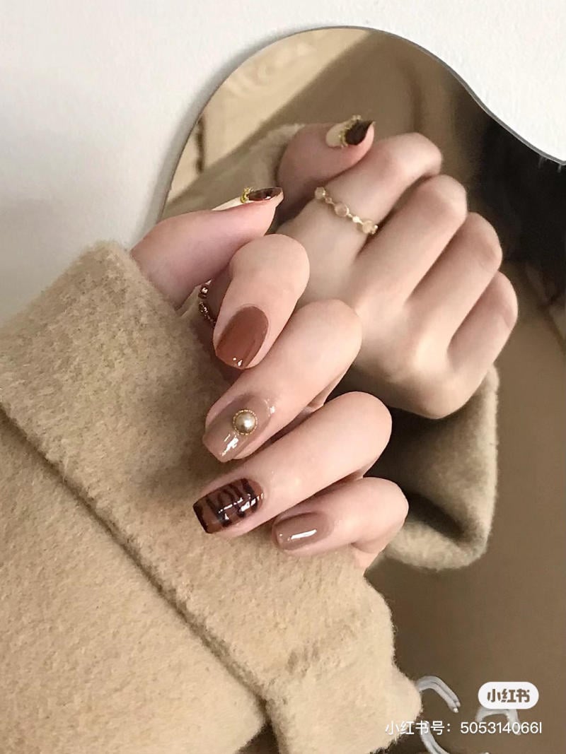 Tay người với móng tay sơn màu nâu, trang trí họa tiết và hạt đính, áo khoác màu be.