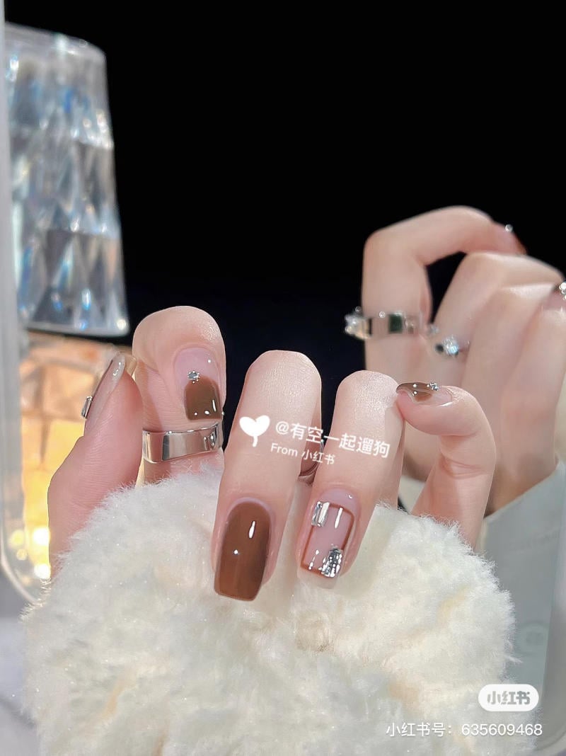 Bàn tay đeo nhẫn với bộ móng tay được sơn màu nâu và trang trí họa tiết lấp lánh.